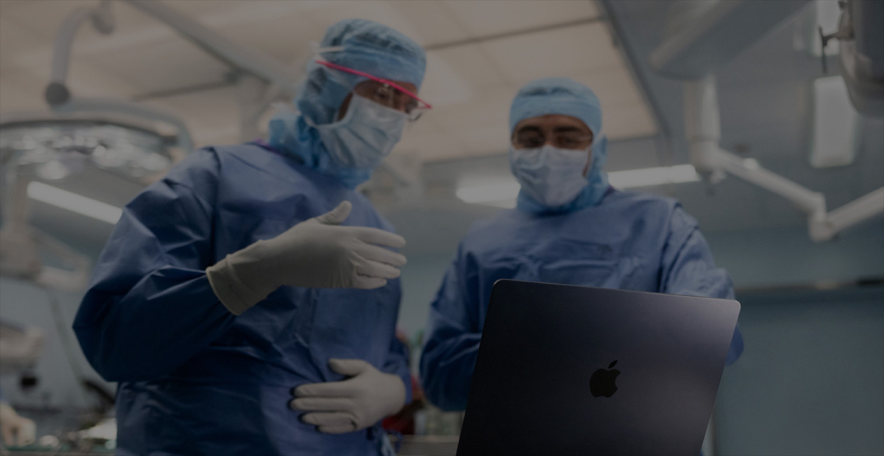 Dois cirurgi?es de uniforme olham para um MacBook Pro na sala de cirurgia.